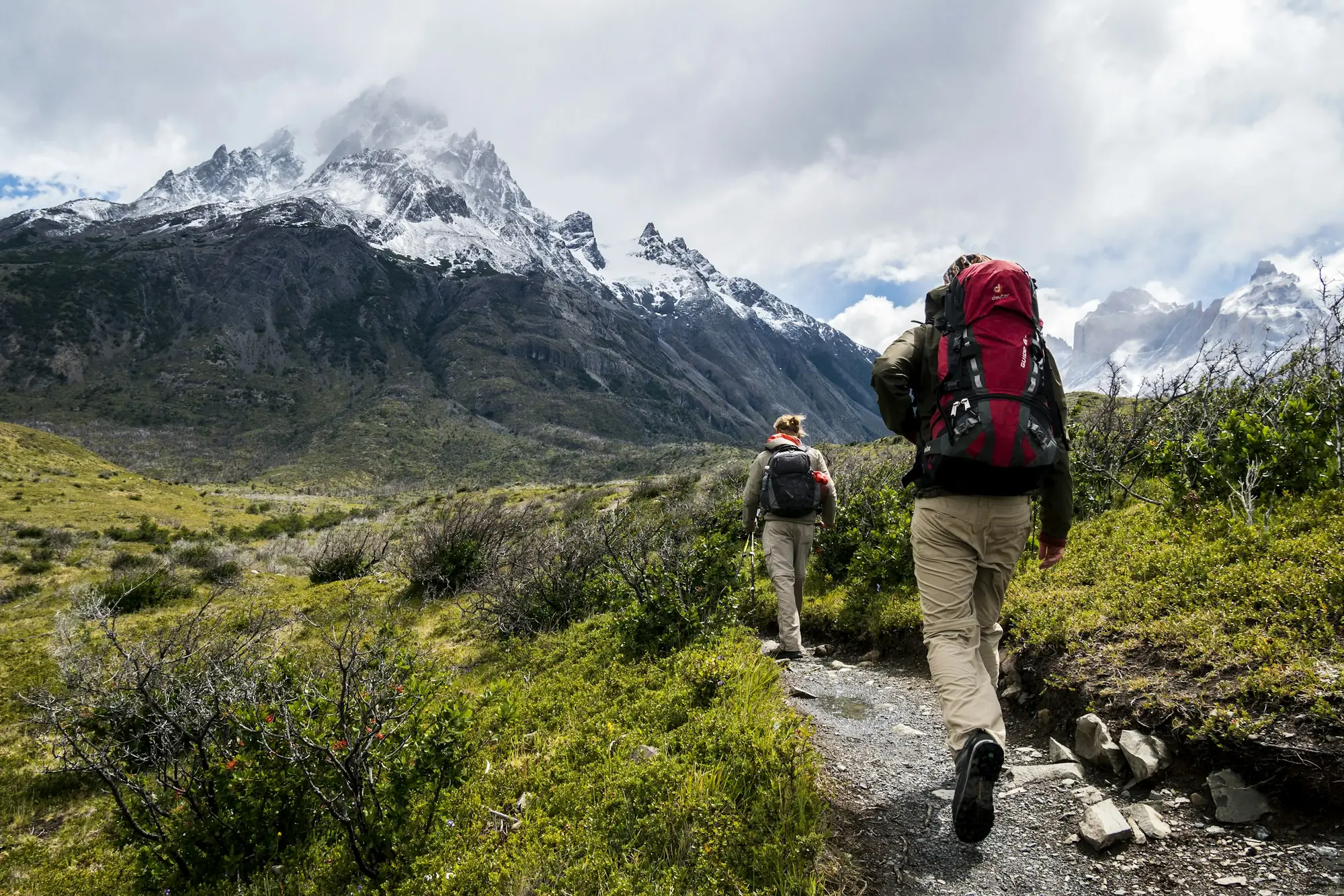 Two hikers walking a rocky trail towards snowcapped peaks
